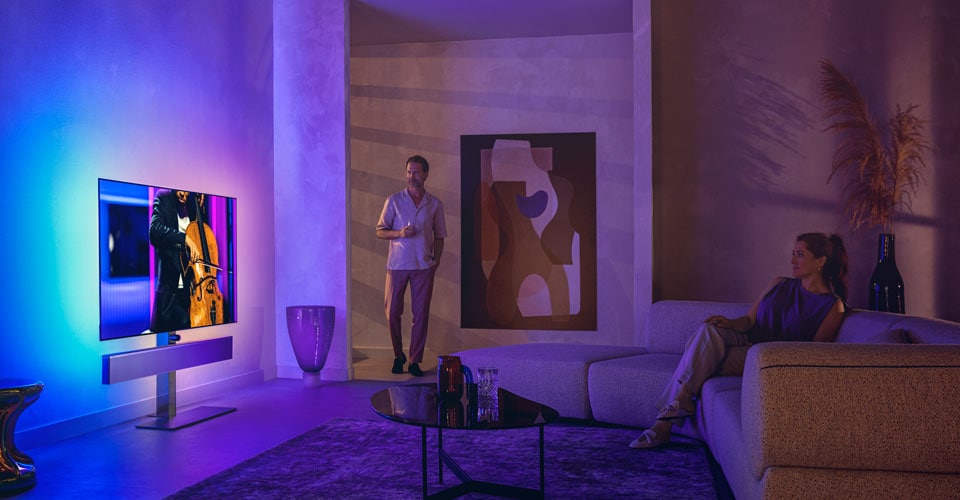Férfi és nő egy stílusos nappaliban, ahol egy csellóelőadást közvetítő Philips Ambilight tévét néznek