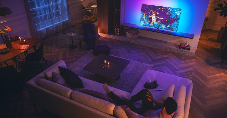 Férfi pihen a kanapén egy halványan megvilágított nappaliban, és Philips Ambilight tévét néz.