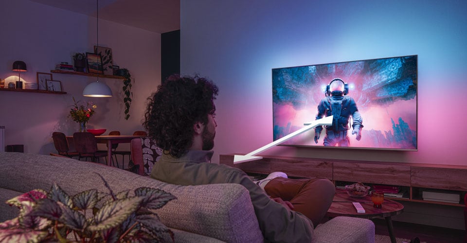 Egy férfi a kanapén ülve néz egy Philips Ambilight tévét, amelyen egy színes űrhajósjelenet látható, egy nyíllal jelezve az optimális nézési távolságot
