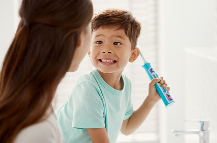 Egy kisfiú grimaszolva édesanyja felé mutatja fogsorát, miközben egy kék Philips Sonicare for Kids gyermek fogkefét tart a kezében. 